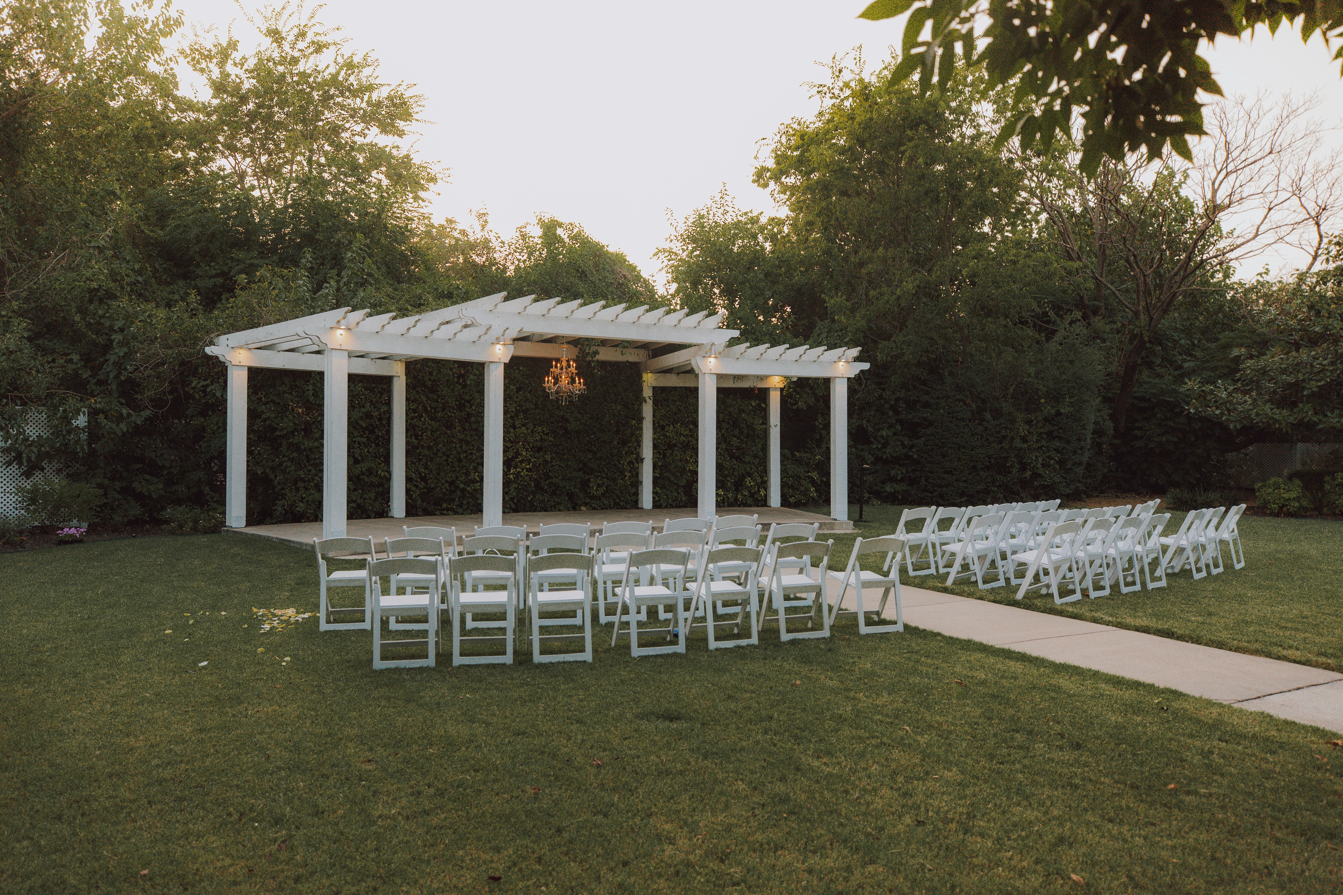 White folding chairs arranged under a pergola with string lights at dusk